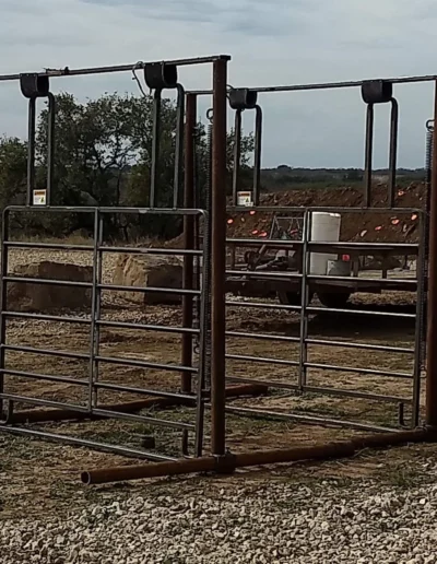 Two metal cattle chutes set on gravel, with a trailer and rocky terrain in the background under a cloudy sky.