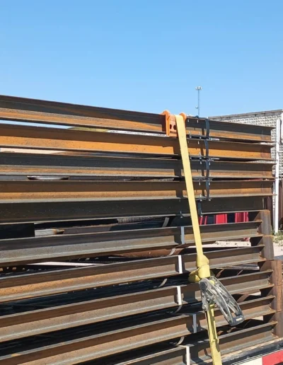 A close-up of a trailer carrying stacked steel framing beams, secured with yellow straps against a clear blue sky.