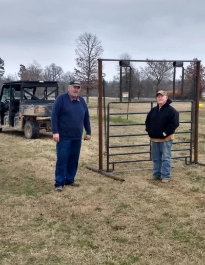 Two men stand beside an open metal gate on a grassy field with trees in the background, while a utility vehicle is parked nearby.